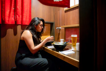 Latina happy woman sits in a private booth enjoying a bowl of ramen and a glass of beer Tokyo, Japan