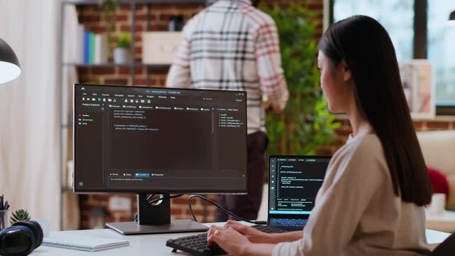 Asian woman is seen typing code for an app development project on her computer, working from a home office. IT specialist engineer focuses on software programming and remote tech work. Camera B.