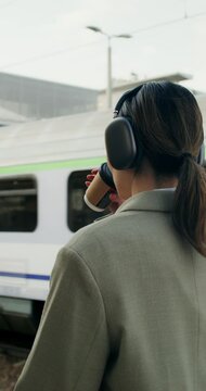 A woman in formal clothes and with headphones on her head uses a mobile phone and drinks coffee from a disposable cup walking along the railway platform