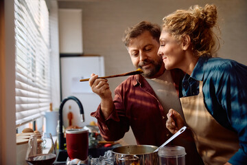 Smiling couple tasting food while preparing Thanksgiving dinner and cooking in kitchen.