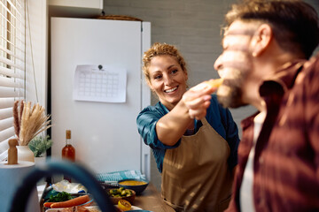 Happy woman feeding her husband and having fun while preparing Thanksgiving dinner in the kitchen.