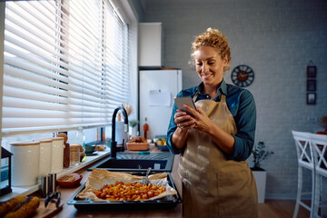 Happy woman texting on cell phone while preparing Thanksgiving meal in kitchen.