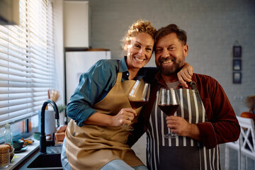 Happy couple drinking wine while cooking in kitchen and looking at camera.