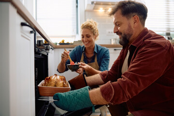 Happy man roasting turkey while making Thanksgiving dinner with his wife in kitchen.