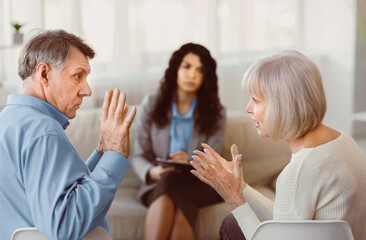 Obraz premium Conflict. Mature couple having fight in therapy or marriage counseling, waving hands, psychologist in the background