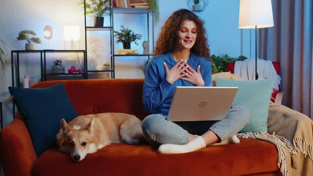 Smiling young woman with curly hair making video webcam conference call while sitting beside corgi dog on sofa in living room at home. Freelancer waving working on laptop during remote work at home.