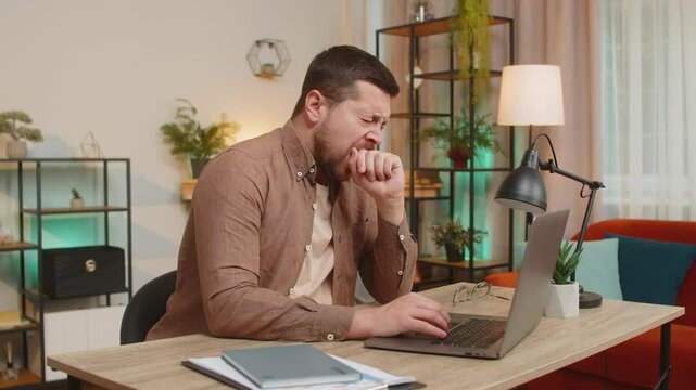 Bored sleepy young businessman working on laptop yawns, sleeping on netbook at home office desk in living room. Exhausted tired Caucasian freelancer workaholic man. Employment, occupation, workless.