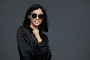 Woman in sunglasses and black shirt, smiling with hands on hips, posing against a gray background