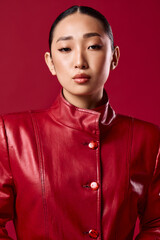 Fashionable asian brunette woman in a red leather jacket poses with hands on hips and head turned to the side, against a red background