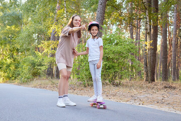 Satisfied woman with brown hair enjoying time outdoor with her daughter on long board looking far at something interesting and pointing away with finger
