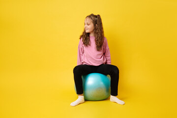 Active little girl with wavy hair engaging with fitness ball gaming looking cheerful away enjoying fun and healthy lifestyle isolated over yellow background