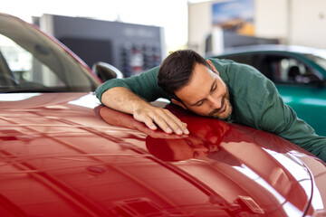 Happy young man hugging his new car in showroom. Satisfied guy with closed eyes embracing the hood of the automobile. Dreaming man lying on car bonnet hugging it.