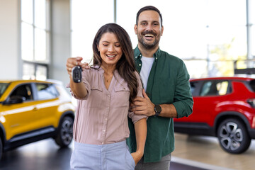 Happy beautiful loving couple buying new car in salon, standing over luxury automobile, hugging and showing key from their new auto, copy space