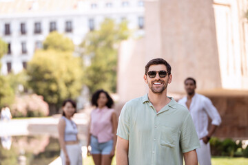 Young man is smiling at the camera with his friends walking behind him during a summer day in the city