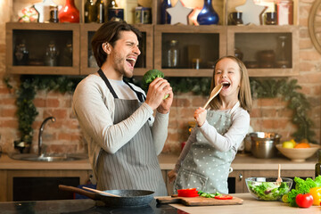 Father and daughter having fun while preparing lunch in kitchen, singing loud, using spatula and broccoli as microphones
