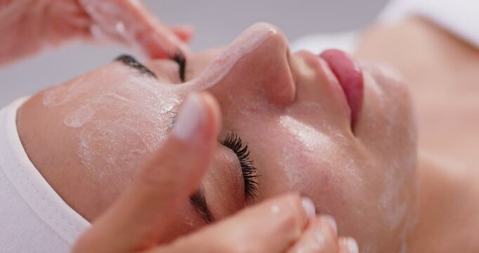 Close up beautician's hands applying anti-aging facial cream on woman client face to prevent wrinkles in spa salon. Skin care, cosmetic procedures for facial care and beauty concept. 4k video.