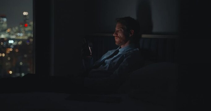 Bored businessman sitting in bedroom overlooking the city at night playing with smartphone or watching videos. Illuminated by phone screen he holds it and scrolls infinitely with tired emotionless