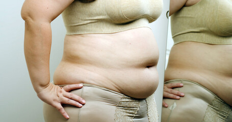 Asian oversize woman in front of a mirror looks at her body. Excess belly fat, Unhealthy lifestyle, Diet concept.