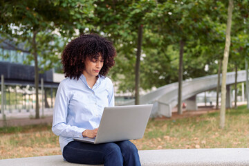 focused female entrepreneur with curly hair sitting on stone bench in park and working remotely on...