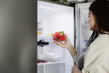 Hungry young pregnant woman standing in front of open fridge, takes strawberry from refrigerator, closeup. Healthy pregnancy eating. Looking for food to have snack, choosing what to have for lunch
