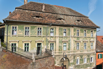 Historic building in Sibiu, Romania, featuring a unique roofline with a circular opening resembling an eye. The building has a green facade with peeling paint.
