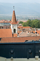 An aerial view of Sibiu, Romania, showcasing a cityscape with red-tiled roofs and a prominent church tower. The tower has a distinctive conical shape with colorful patterns and a clock.