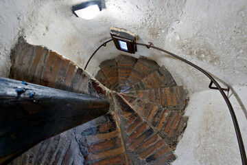 A spiral staircase with worn brick steps and a curved metal railing. The walls are whitewashed, with a small window casting light on the stairs. A support beam is visible on the left side of the image