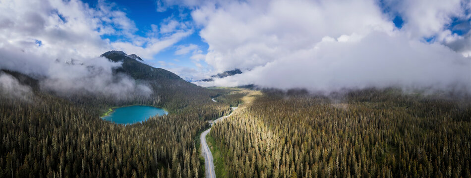 Aerial view of a picturesque lake surrounded by lush forest and a winding road under a blue sky with clouds, Agassiz, Canada.