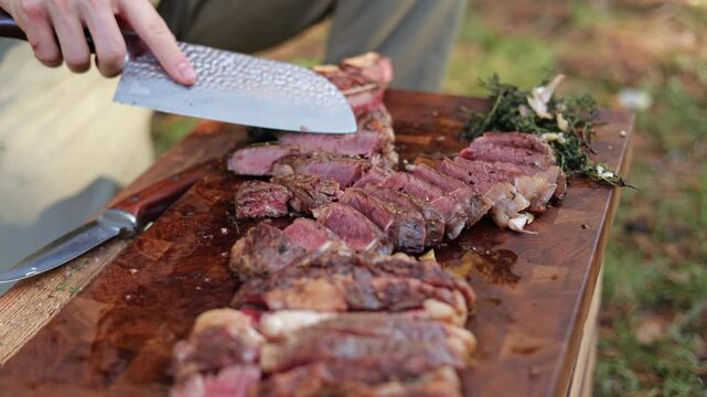 Close up of person slicing medium rare grilled steak on wooden cutting board outdoors. Juicy rib eye steak sliced with knife on butcher block.