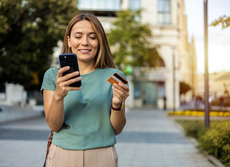 Happy young Caucasian woman uses her smartphone and credit card to shop online while walking outdoors in the city. Smiling woman walking in city with mobile phone and credit card looking at camera.