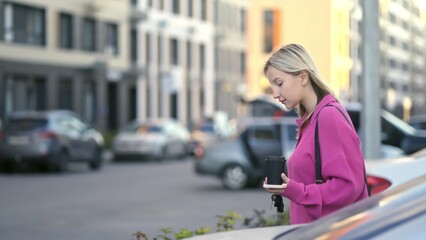 Naklejka premium A young woman dressed in a pink sweater gazes down at her smartphone, surrounded by a vibrant urban backdrop