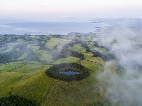 Aerial view of serene Lagoa Pau Pique surrounded by lush green fields and trees, Santo Antonio, Portugal.