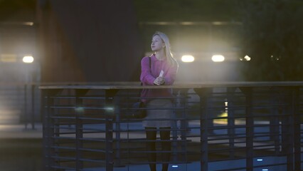 A woman stands thoughtfully by a railing at night, deeply lost in contemplation against the vibrant...