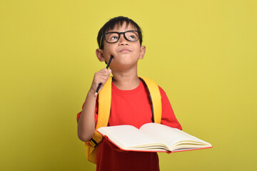 Portrait of an Asian boy with backpack holding pencil and open book posing thinking solving problem...