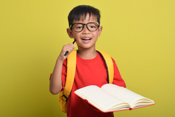 Portrait of an Asian boy with backpack holding pencil and open book posing thinking solving problem isolated on yellow background. Time to study. Back to school