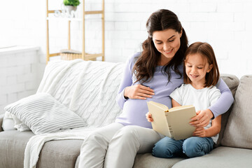 Family time. Preschool girl and her expecting mom reading exciting book at home, copy space