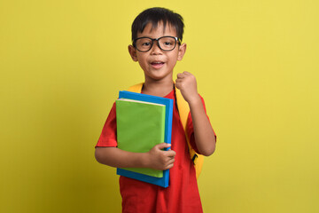 Cute and cheerful Asian boy smiling wearing backpack holding book gesturing showing victory sign isolated on yellow background. Time to study. Back to school