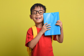 Asian boy holding blue book in his hand isolated on yellow background. A little boy excited for school