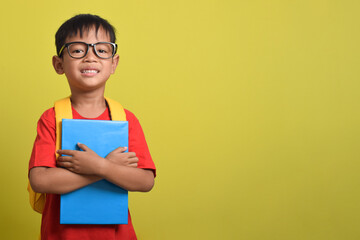 A smiling Asian boy holding a book isolated on a yellow background. A little boy getting ready for school
