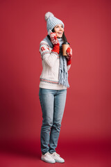 Fototapeta premium Cheerful woman in cozy Christmas sweater talking on smartphone and standing with warm drink in a festive studio