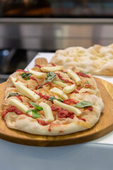 Close-up side view of fresh baked pizza with cheese, basil leaves and tomato paste on wooden board on professional kitchen table in pizzeria. Soft focus. Copy space. Cooking class. Food business theme