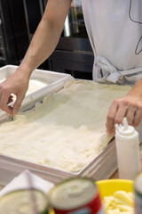 Close-up side view of male cook hands preparing thin rectangular pizza dough on professional kitchen table in pizzeria or italian restaurant. Soft focus. Copy space. Food service business theme.