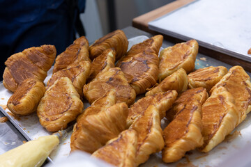 Close-up side view of fresh baked brown croissants lying upside down on baking tray before filling with cream on professional kitchen table. Soft focus. Copy space. Food preparation theme.
