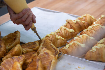 Close-up side view of cook decorating fresh baked croissant with custard using icing bag in professional bakery kitchen. Soft focus. Pastry preparation  theme.