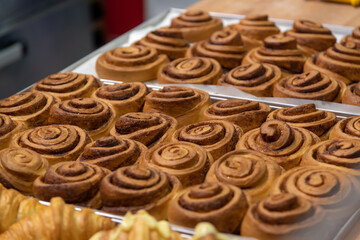 Close-up side view of fresh baked brown cinnamon buns and croissants on baking trays on professional kitchen table in bakery. Soft focus. Copy space. Pastry business background theme.