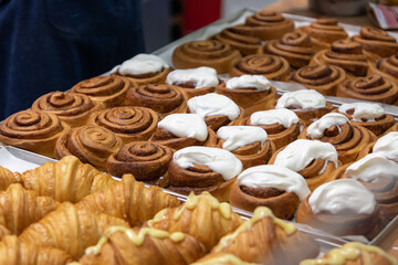Close-up side view of various fresh baked puff pastry items (croissants with different fillings and cinnamon rolls) on baking trays on professional kitchen table. Soft focus. Food background.