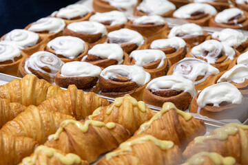 Close-up side view of various fresh baked puff pastry items (croissants with different fillings and cinnamon rolls) on baking trays on professional kitchen table. Soft focus. Food background.
