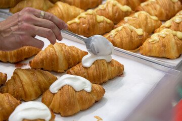 Close-up side view of baker putting whipped cream on top of fresh baked brown traditional croissants using spoon on professional kitchen table. Soft focus. Food preparation and decoration theme.
