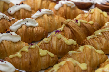 Close-up side view of various fresh baked puff pastry items (croissants with different fillings with edible flowers and cinnamon rolls) on baking tray on professional kitchen table. Food background.