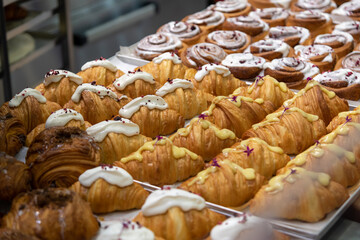 Close-up side view of various fresh baked puff pastry items (croissants with different fillings with edible flowers and cinnamon rolls) on baking tray on professional kitchen table. Food background.